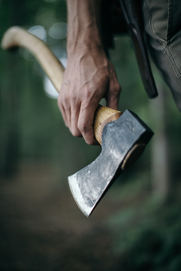 A hand clasps a long axe in front of a blurred forrest background.