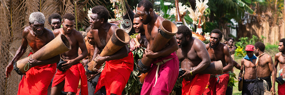 Tribespeople bang their drums, wearing bright red shorts