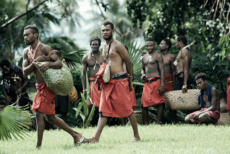 A group of native tribesmen wearing traditional red garments walk across the scene.